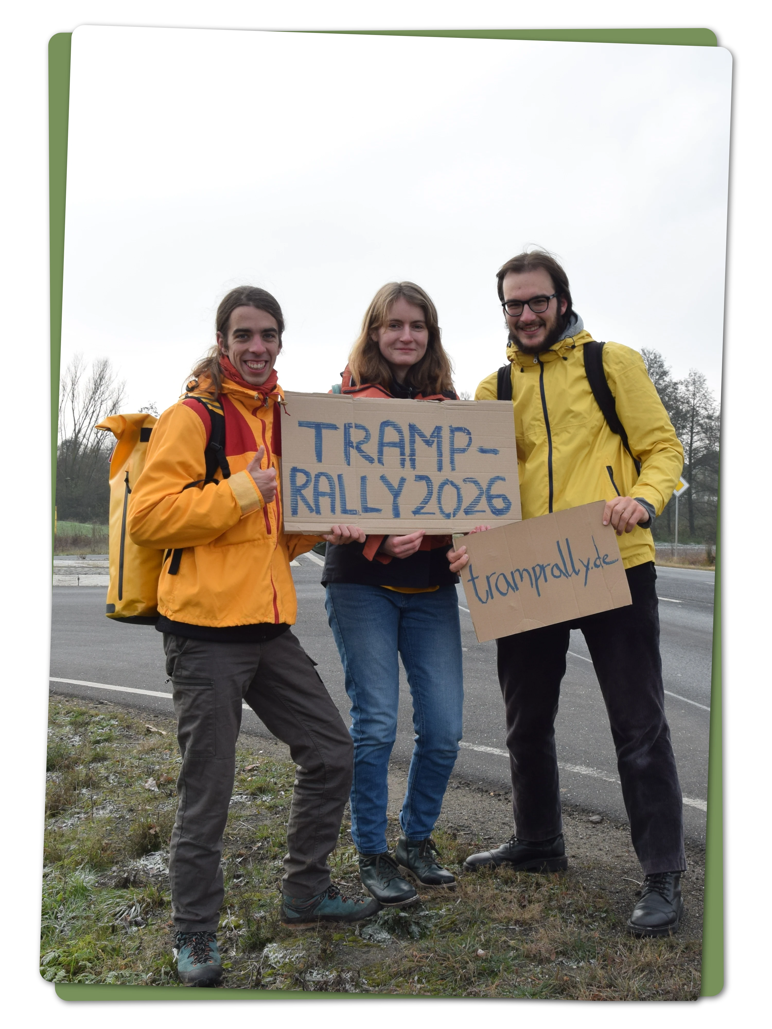 Two hitchhikers are standing by a highway an holding up handwritten signs reading "Tramprally 2025, sign up now"