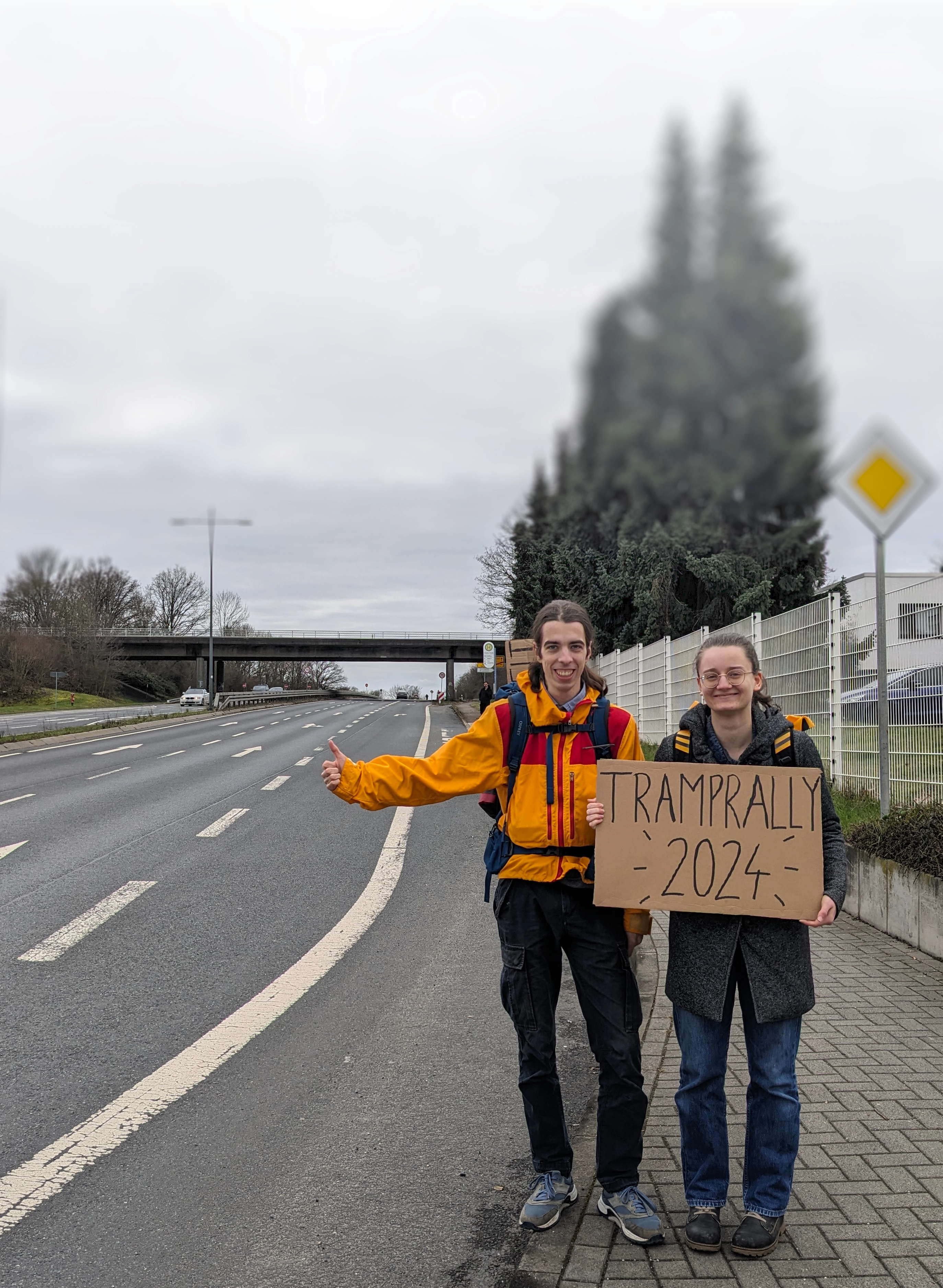 Zwei Tramper stehen mit ausgestrecktem Daumen am Straßenrand und halten ein handgeschriebenes Pappschild mit der Aufschrift "Tramprally 2024"