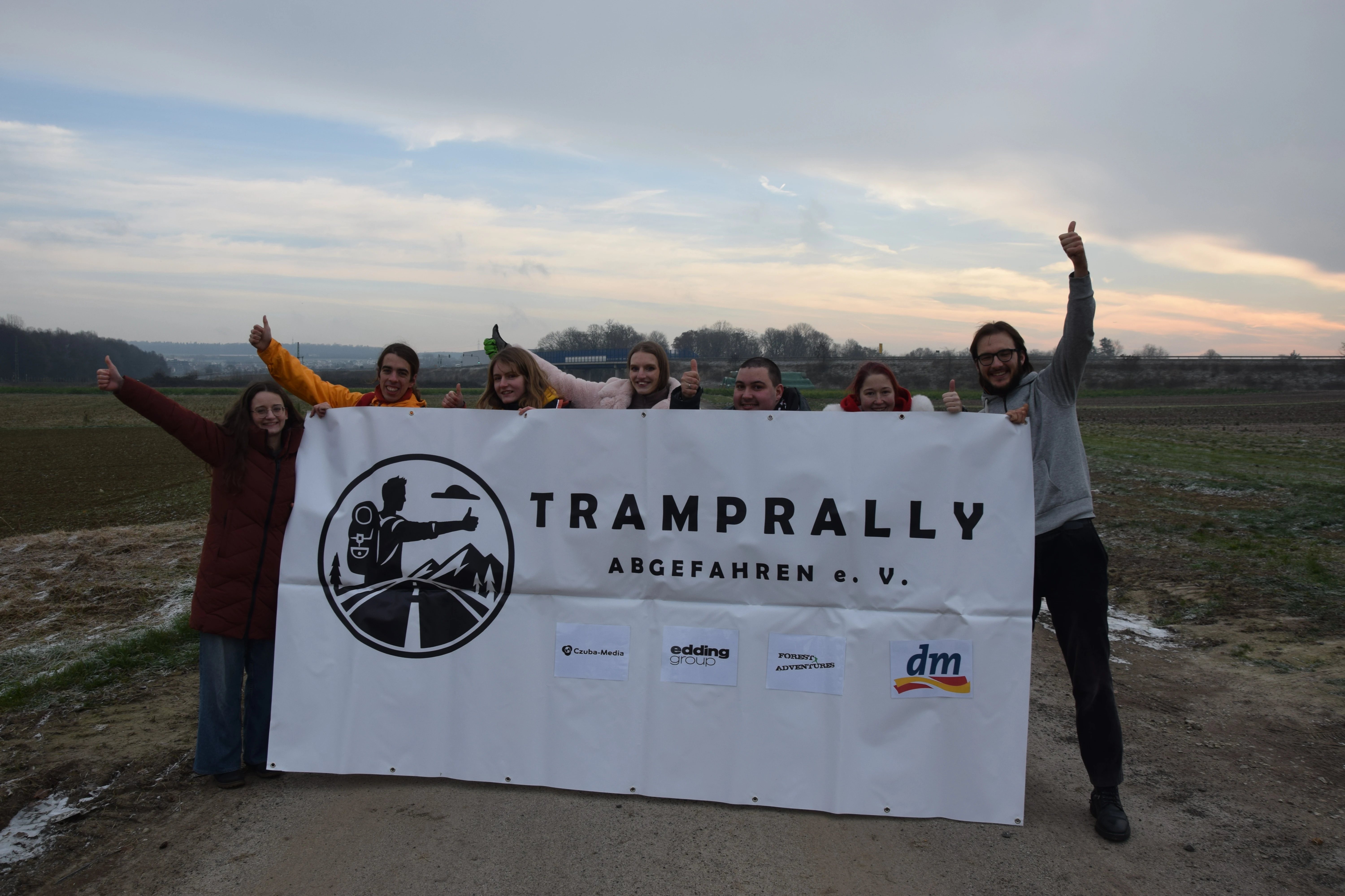 Group photo of the team members holding a large Tramprally banner.
Everyone is giving a thumbs up gesture.
The backdrop is a field in front of a highway.
