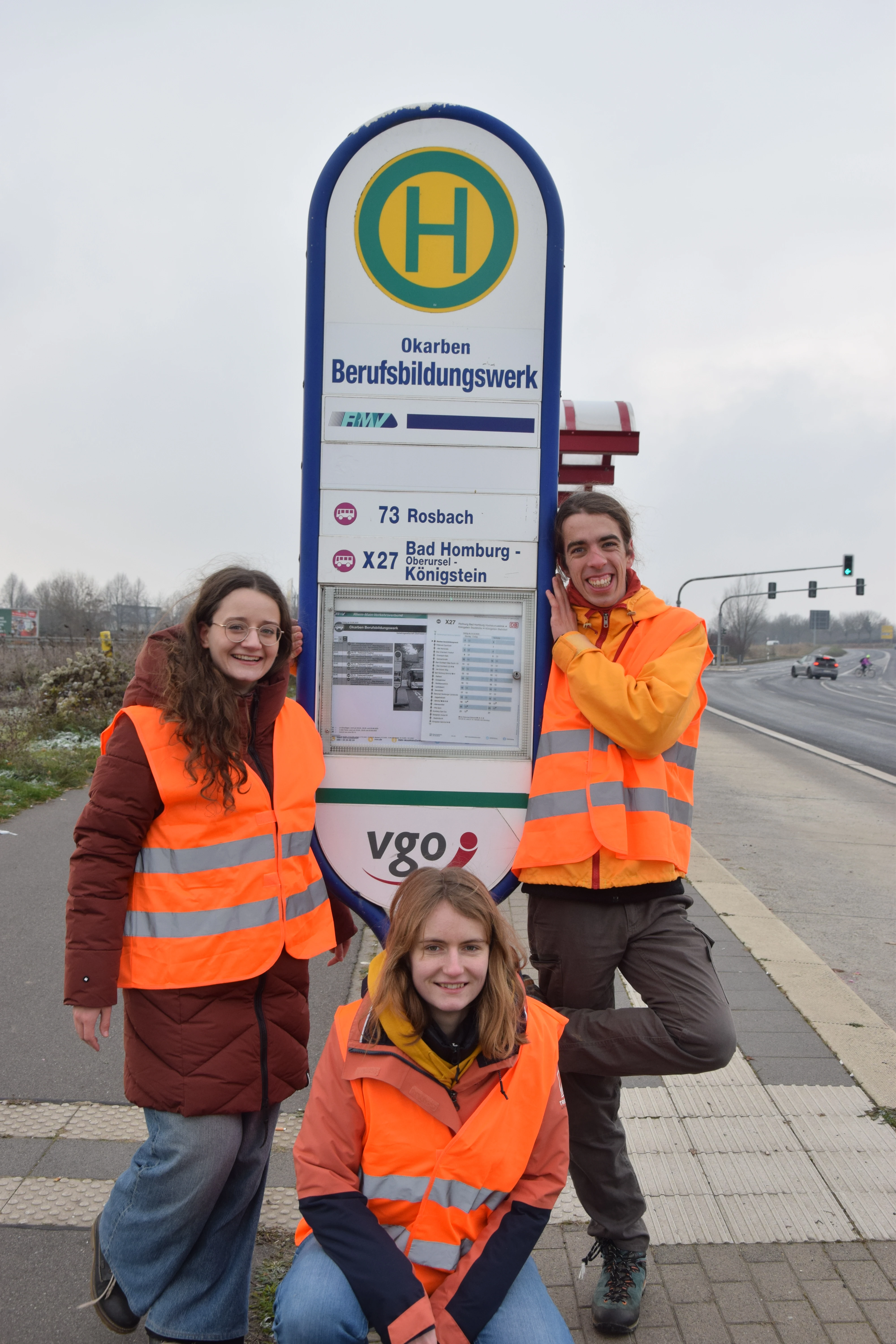 Three members of the team are standing at a bus stop. They are wearing bright orange high-viz vests which contrast against the dull gray sky. They are all smiling.
