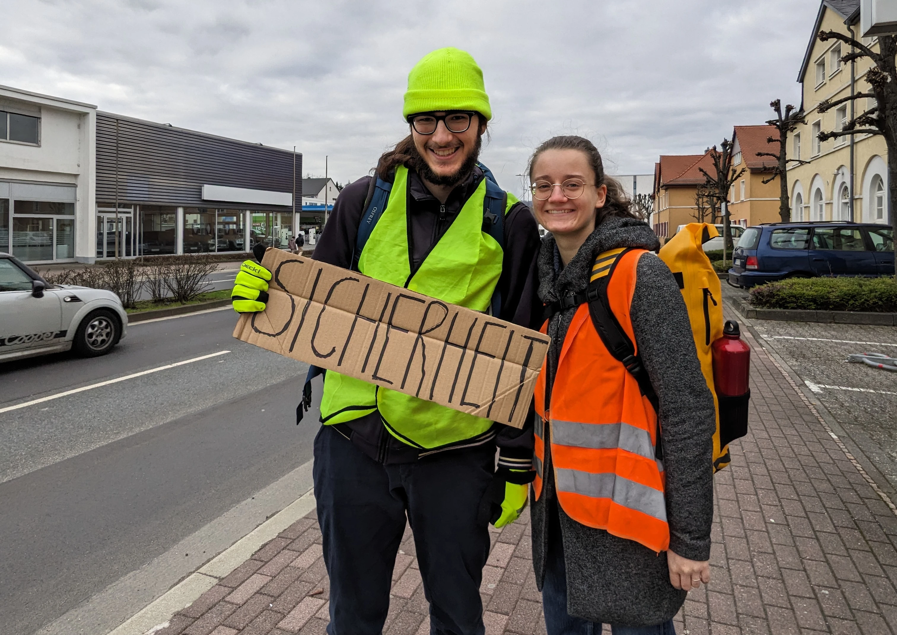 Zwei Menschen in Warnwesten stehen am Straßenrand und halten ein handgeschriebenes Schild mit der Aufschrift "Sicherheit"