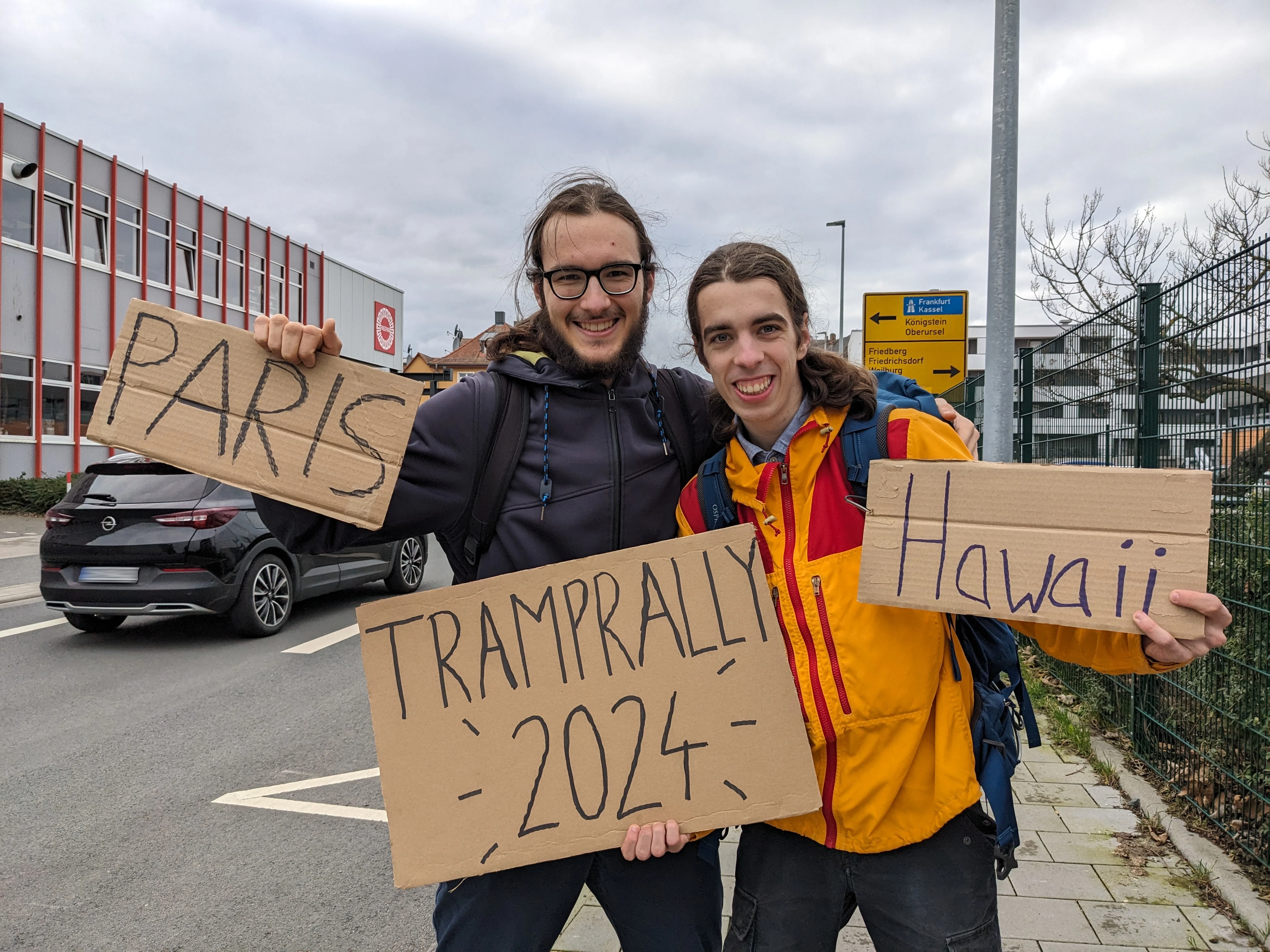 Zwei lachende Tramper stehen am Straßenrand und halten handgeschriebene Pappschilder mit den Aufschriften "Paris" und "Hawaii"